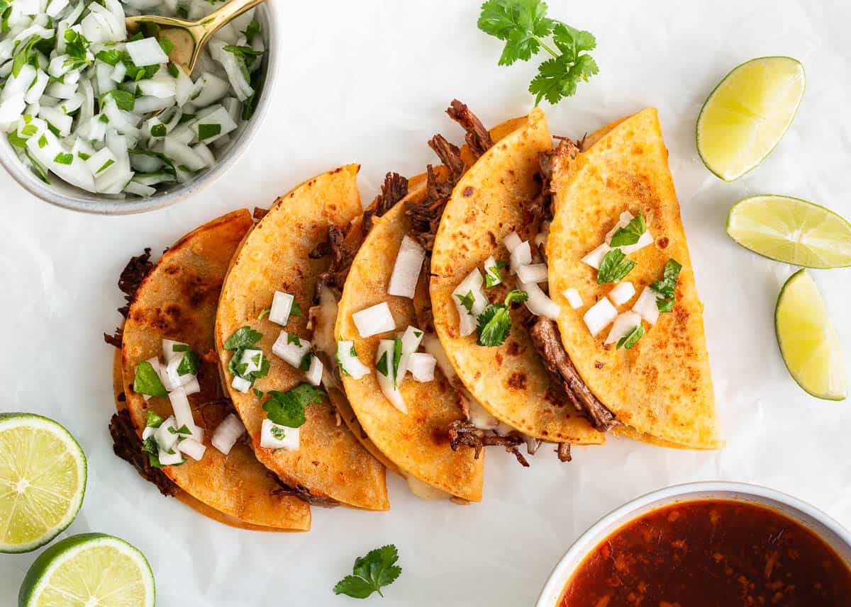 These birria tacos are made with corn tortillas dipped in a flavorful broth, filled with tender beef and cheese and then pan fried until crispy quesabirria perfection! Five assembled birria tacos on parchment paper on the counter.
