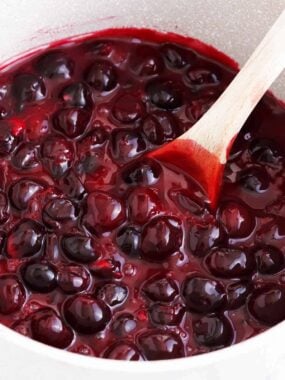 A close up of cherry pie filling in a pot with a wooden spoon.