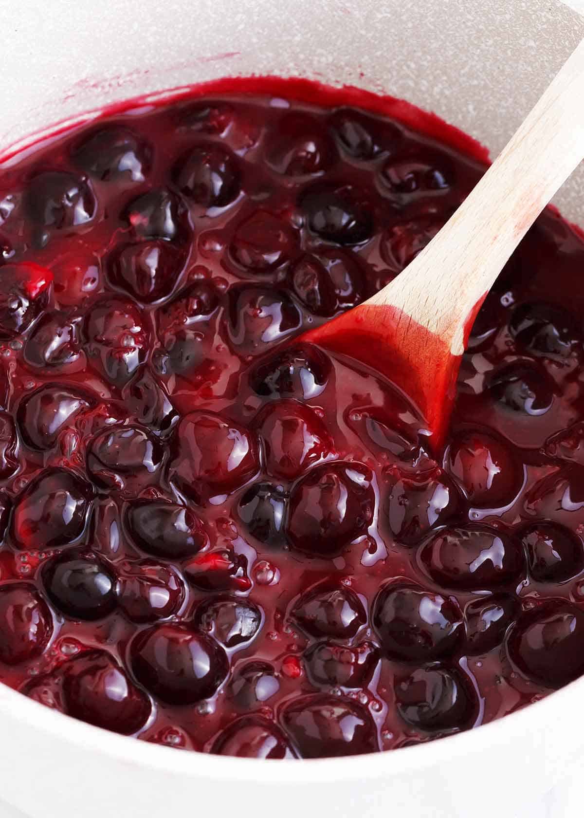 A close up of cherry pie filling in a pot with a wooden spoon.