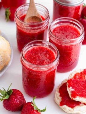 Four jars of strawberry jam on the counter with a biscuit split open to the side spread with jam.