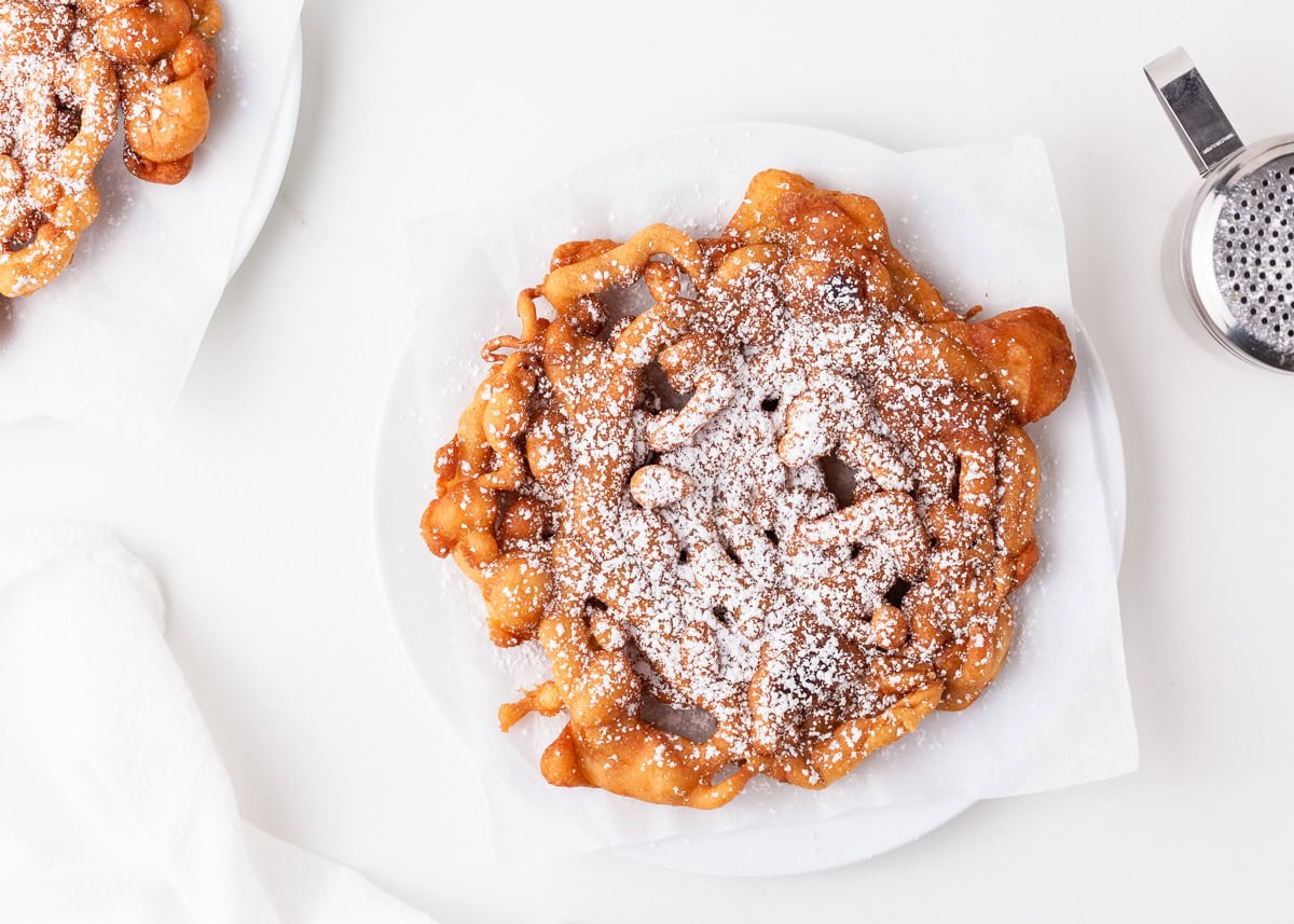 Homemade funnel cake on a white plate dusted with powdered sugar, lacy spirals visible.