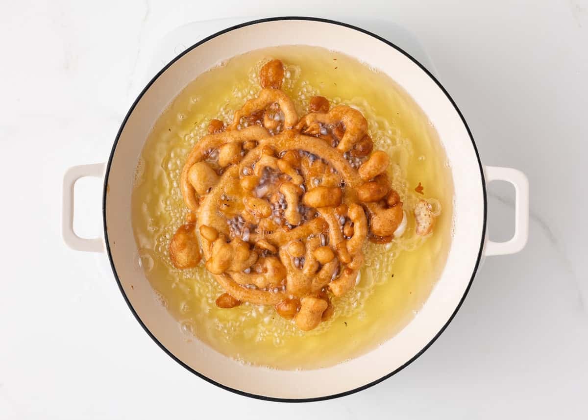 Funnel cake frying in hot oil in a white Dutch oven, edges turning golden brown and crisp.