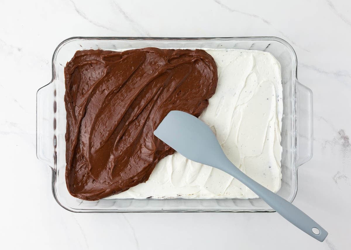 Chocolate pudding layer being spread over the cream layer in a glass baking dish.