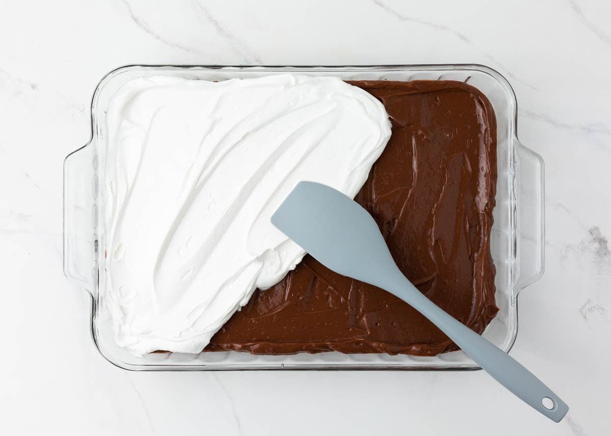 Cool Whip topping being spread over the chocolate pudding layer with a spatula.