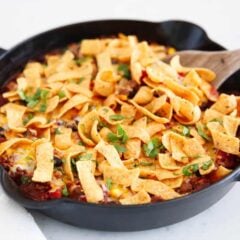 Frito pie in a cast iron skillet topped with melted cheese, crunchy Fritos, and fresh cilantro, with a wooden spoon resting on the side.