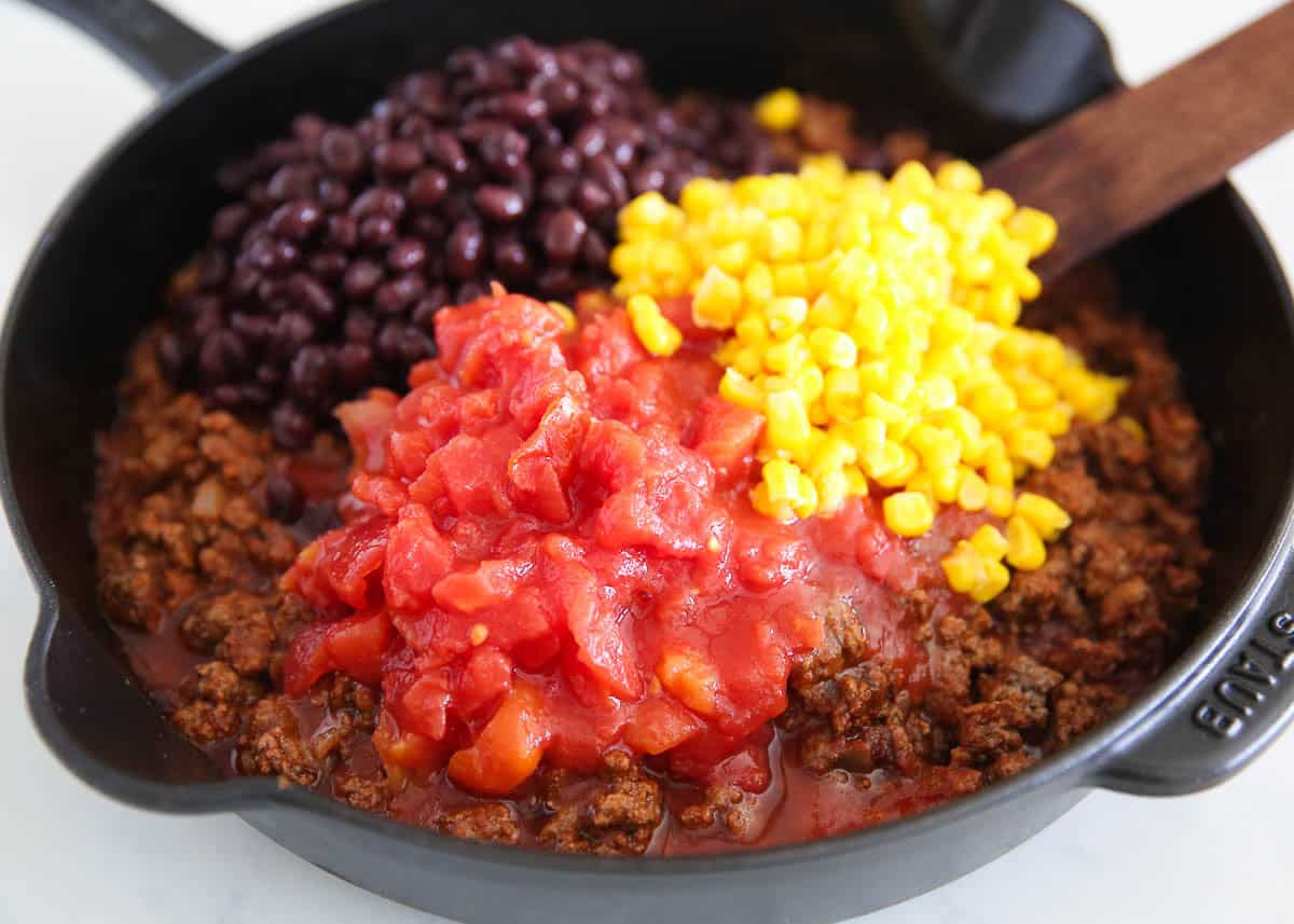 Browned ground beef in a cast iron skillet topped with black beans, corn, and diced tomatoes before stirring together for easy Frito pie.