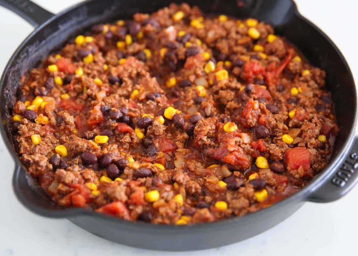 Frito pie filling with seasoned ground beef, black beans, corn, and diced tomatoes simmering in a black cast iron skillet.