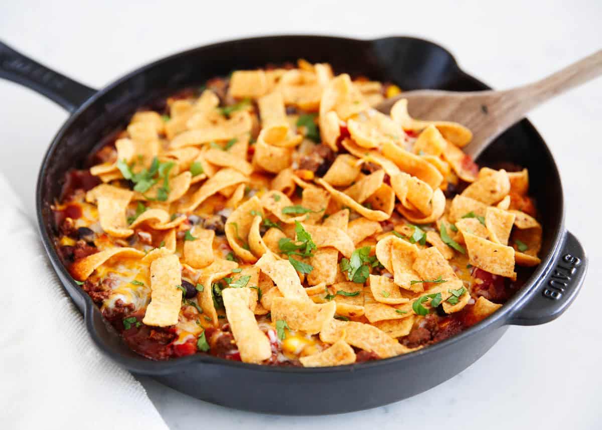 Frito pie in a cast iron skillet topped with melted cheese, crunchy Fritos, and fresh cilantro, with a wooden spoon resting on the side.