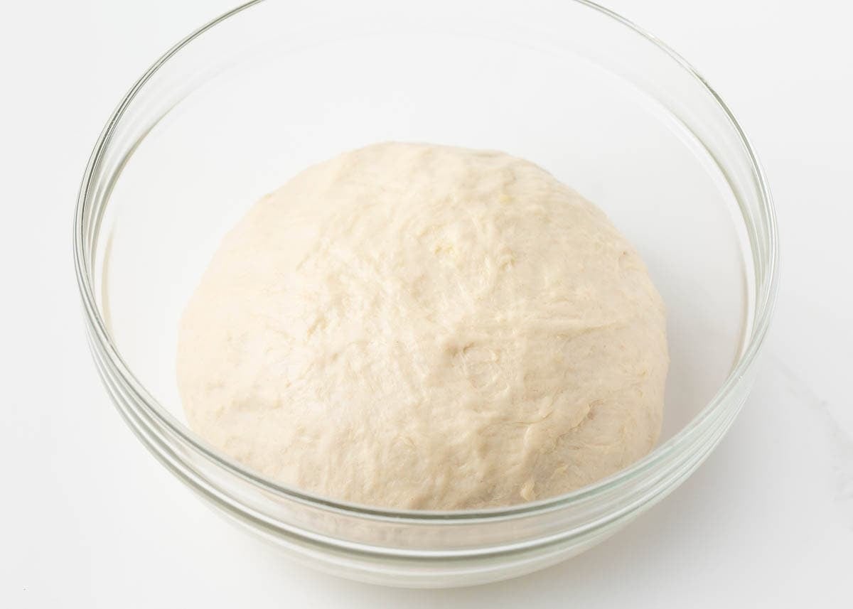 Smooth ball of homemade pizza dough resting in a glass bowl before shaping.
