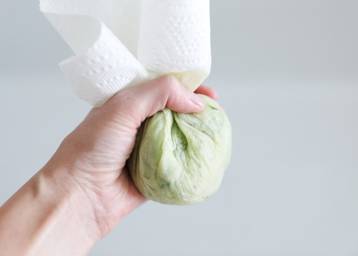 Hands squeezing shredded zucchini in a paper towel over a bowl for banana zucchini muffins.