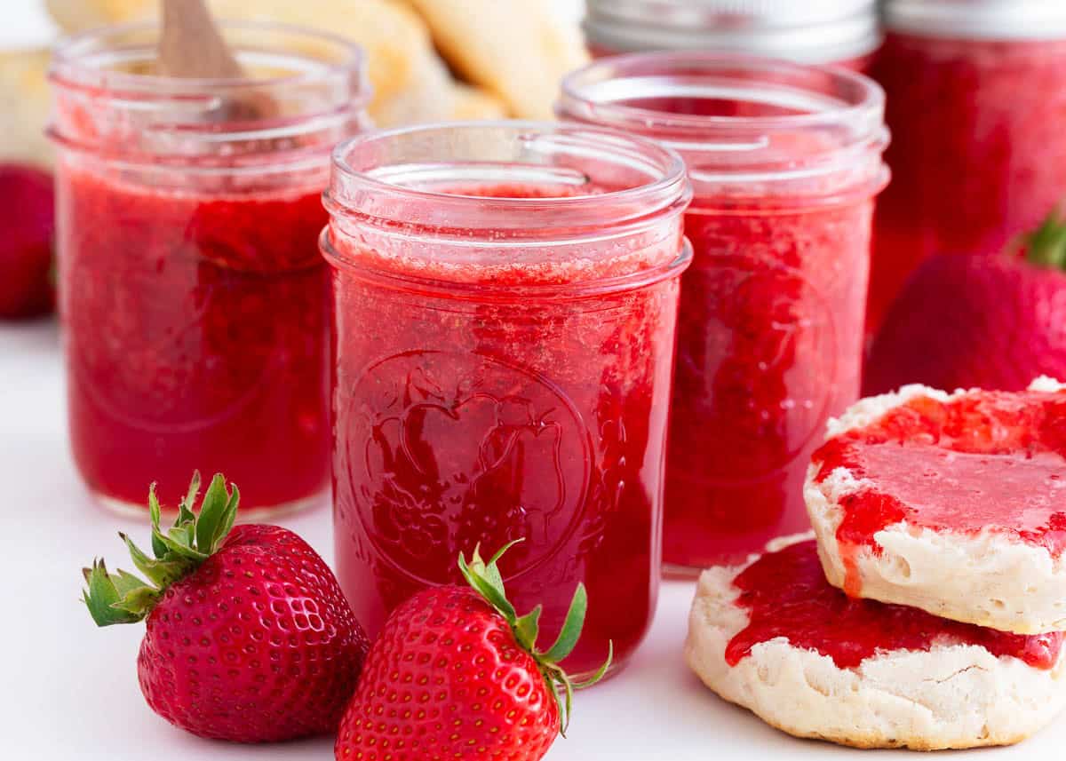 The best homemade strawberry jam that's easy to make with only four ingredients! Full of fresh sweet berry flavor, it beats out store bought any day. Jars of homemade strawberry jam with fresh strawberries and a split biscuit spread with jam.