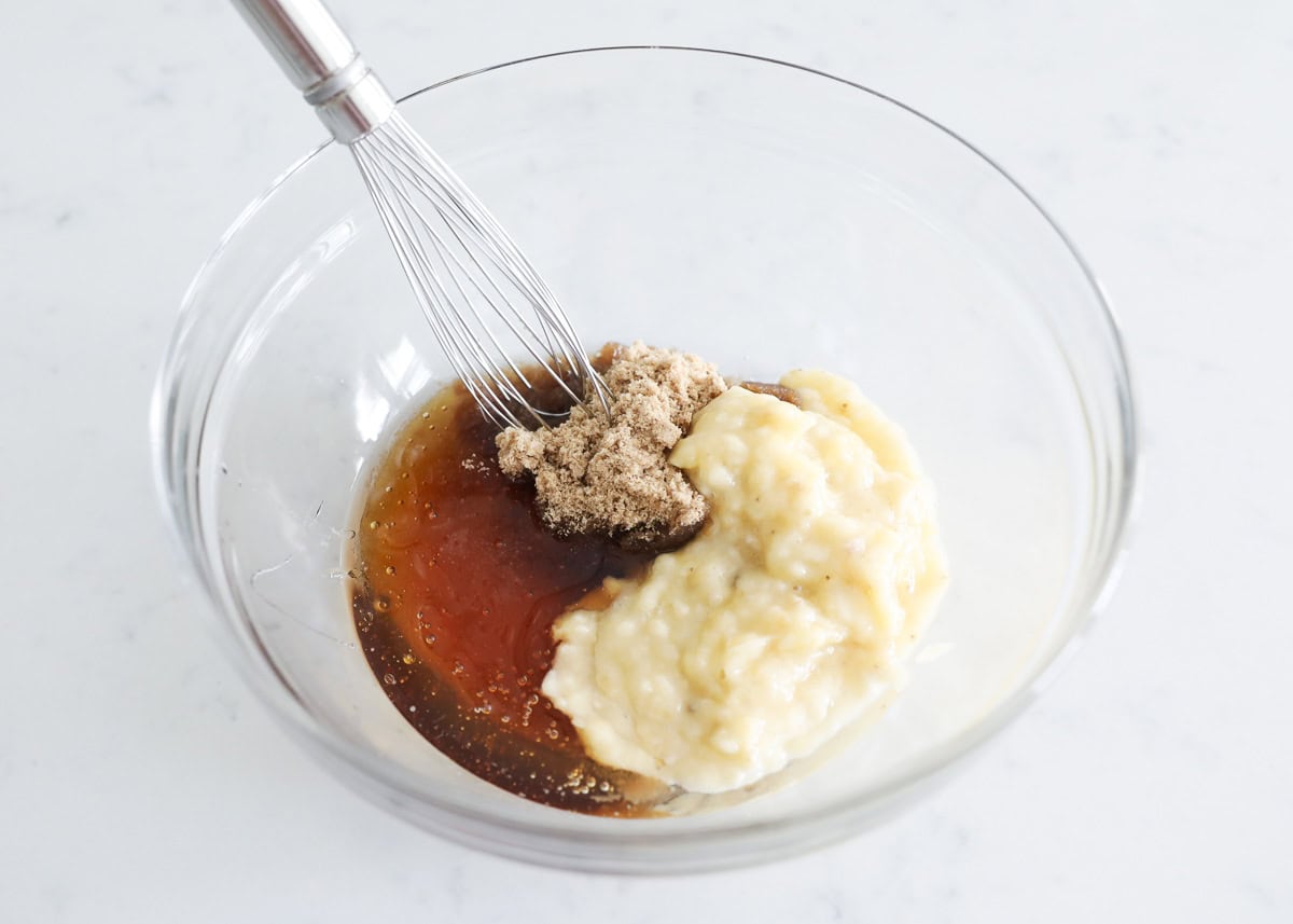 Wet ingredients—mashed banana, honey, brown sugar, eggs, and oil—being whisked together in a large glass bowl.

