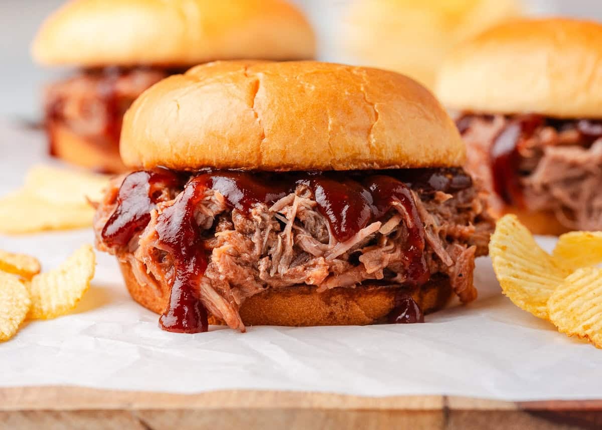 Close-up of BBQ pulled pork sandwiches on soft buns with juicy shredded pork and barbecue sauce, served with potato chips.
