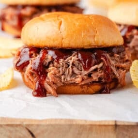 BBQ pulled pork sandwich on a soft bun topped with tangy barbecue sauce, served on parchment with potato chips.