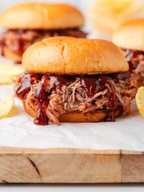 BBQ pulled pork sandwich on a soft bun topped with tangy barbecue sauce, served on parchment with potato chips.