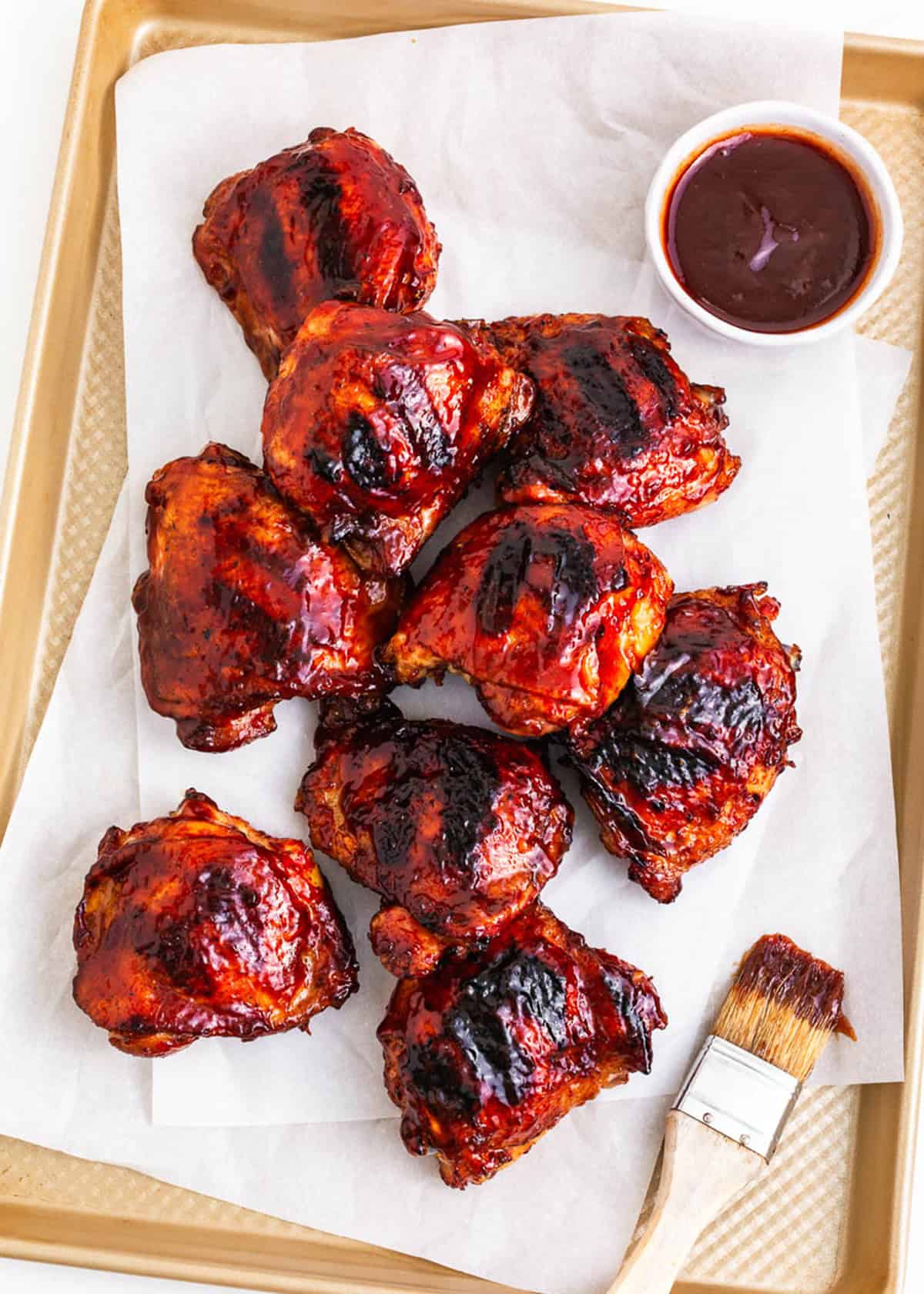 Grilled BBQ chicken on a paper lined baking sheet with bowl of sauce and basting brush.