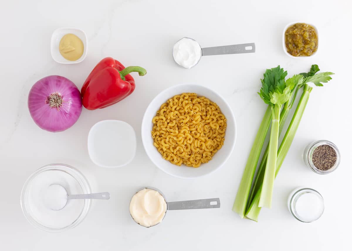 Elbow macaroni, mayo, sour cream, celery, red onion, red bell pepper, Dijon mustard, sweet pickle relish, salt and pepper on the counter.