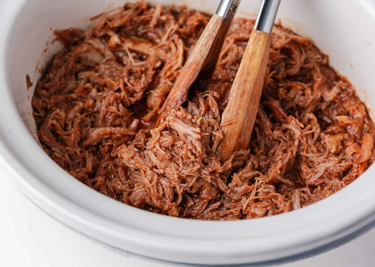 Shredded BBQ pulled pork in a slow cooker being mixed with sauce using tongs.