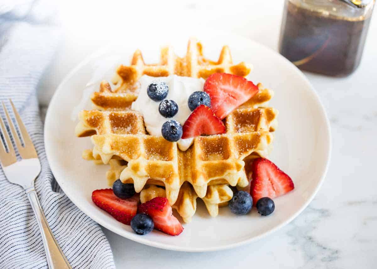 Belgian waffle topped with fruit and whipped cream on a white plate.