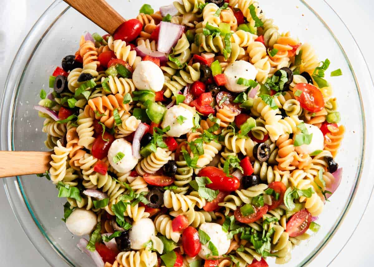 Pasta salad with Italian dressing in a glass bowl with wooden serving utensils. 