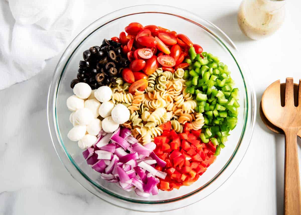 Rotini pasta, green and red bell pepper, mozzarella balls, red onion cherry tomatoes and olives in a bowl with Italian dressing.