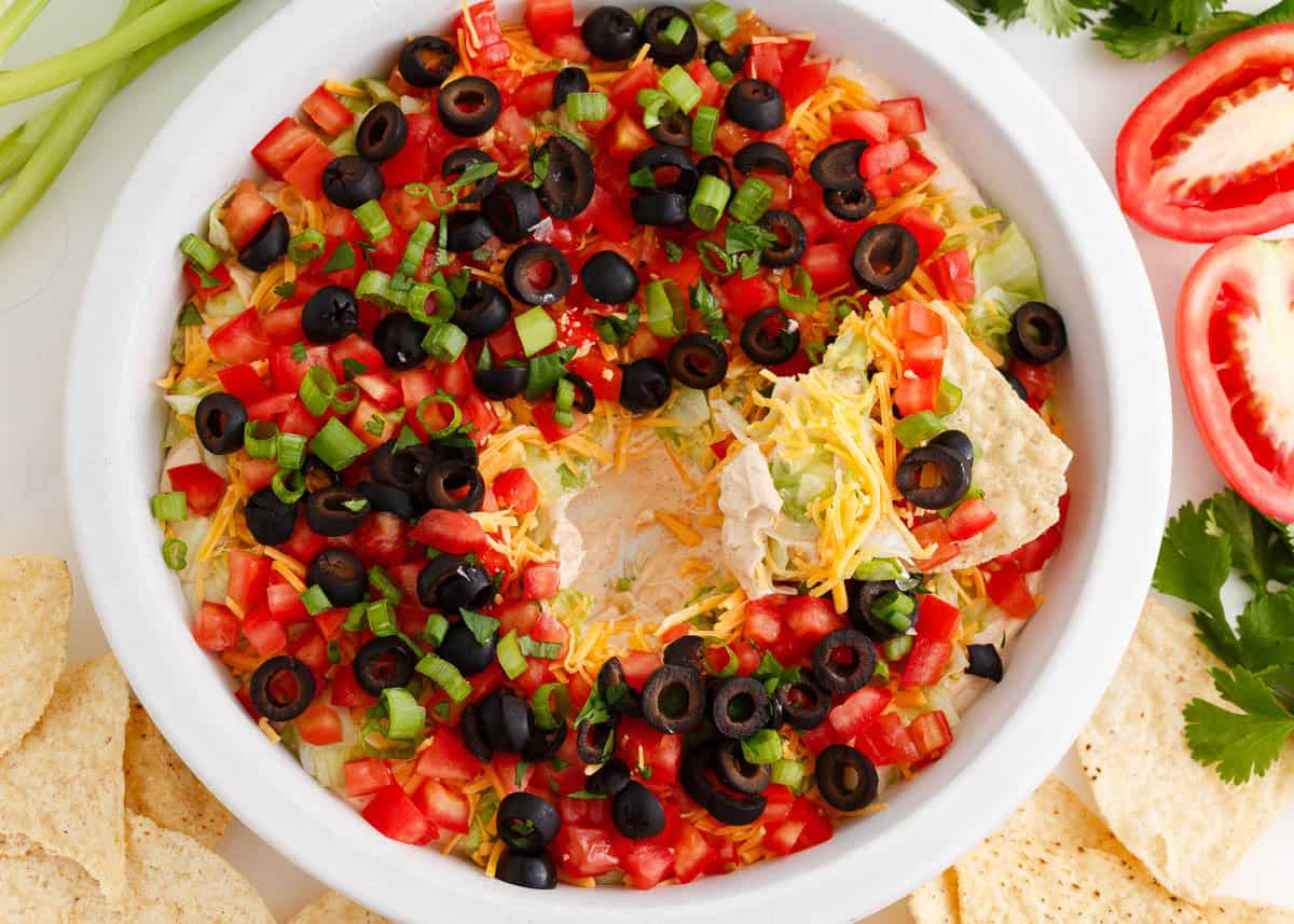 "Taco dip with cream cheese in a white dish with a scoop removed showing the layers, served with tortilla chips.
