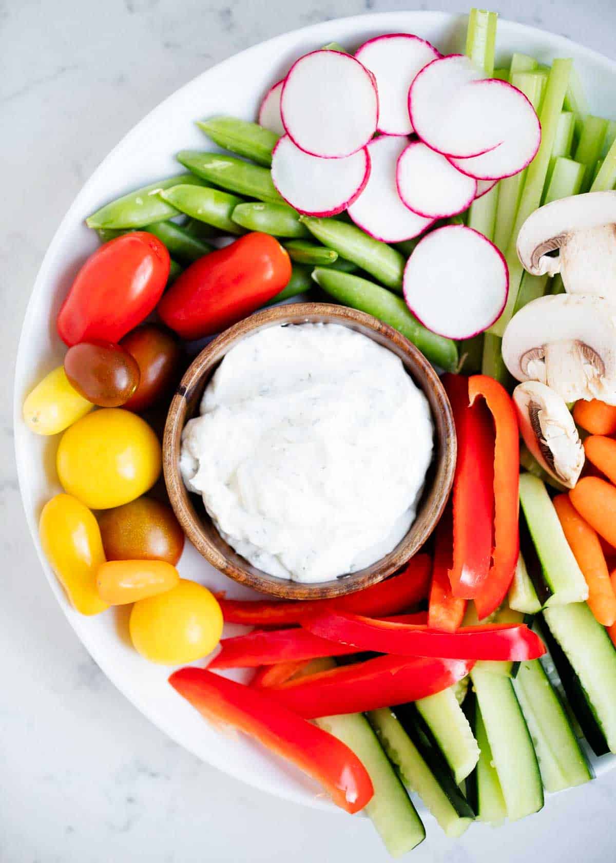 Dill dip with fresh cut veggies around it on a white plate. 