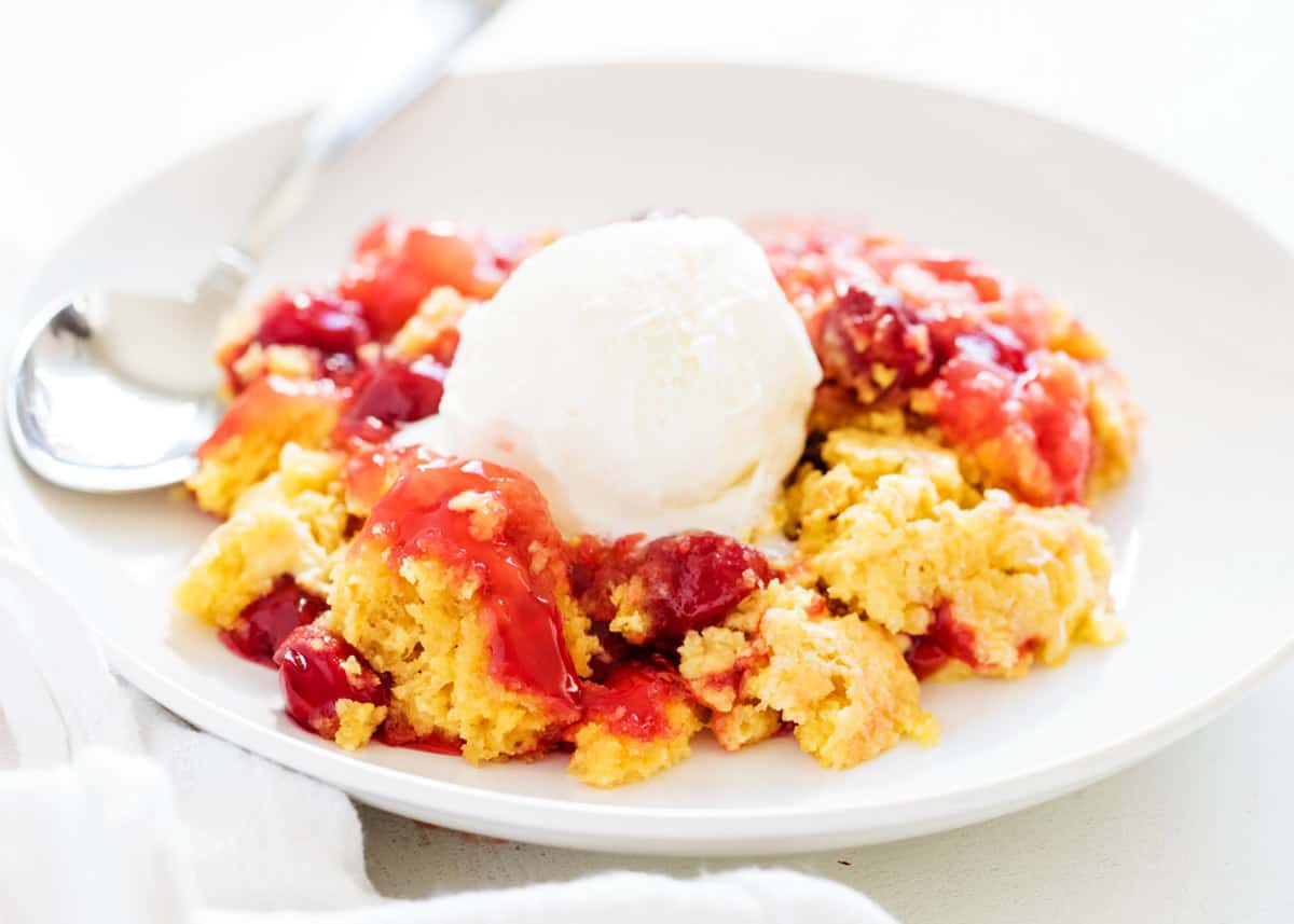 This easy dump cake can be made 4 different ways and calls for just 4 simple ingredients. Such a classic dessert and the perfect recipe to have in your back pocket! Dump cake with ice cream in a white bowl with spoon.