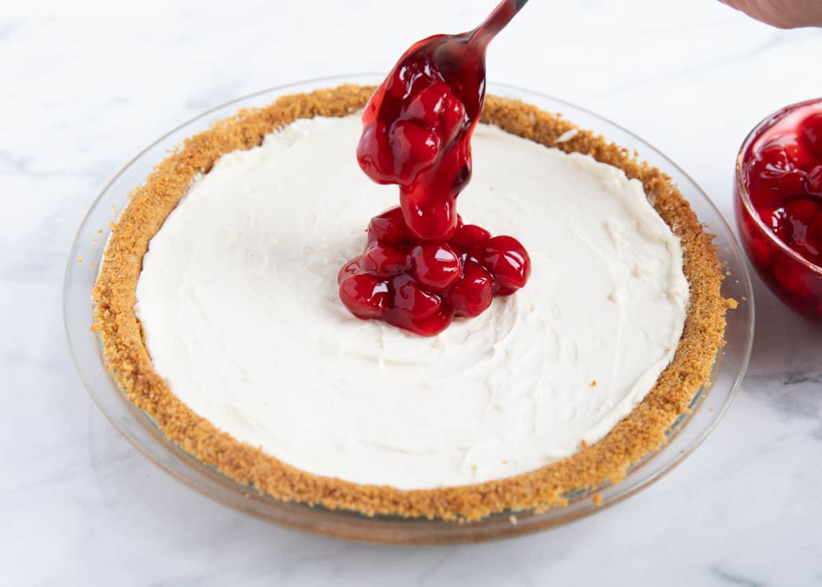 No bake cheesecake with cherrie pie filling being spooned on top of it. 