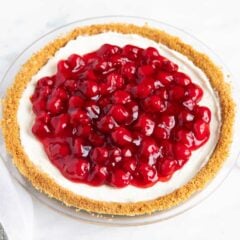 No-bake cheesecake in pie dish on the counter topped with cherry filling with a pie server nearby.