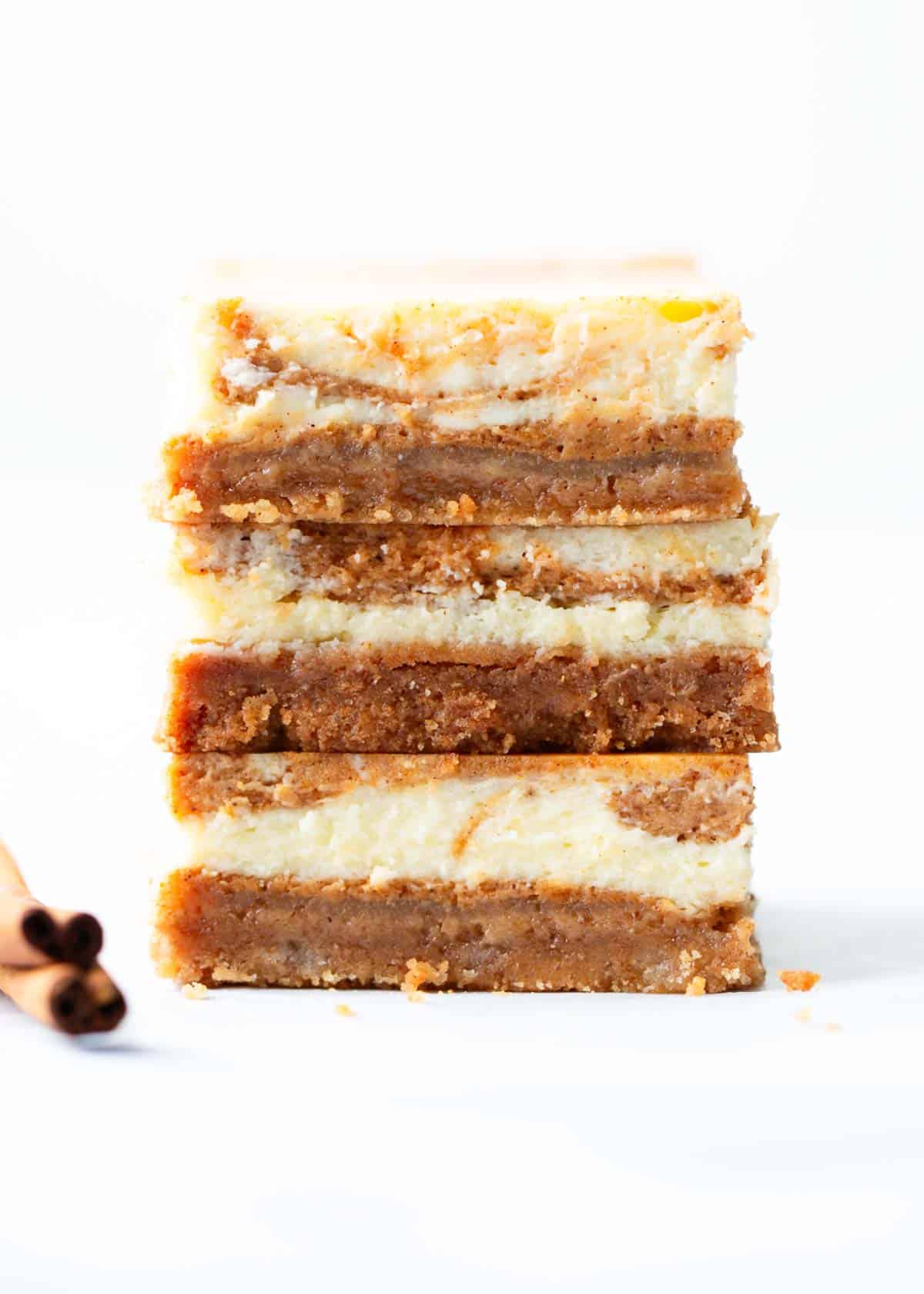 Made with a sweet graham cracker crust and topped with a luscious and creamy cheesecake and pumpkin filling, these pumpkin cheesecake bars are a lighter and fluffier cheesecake version of my popular pumpkin pie bars! Stack of pumpkin cheesecake bars on a white counter.