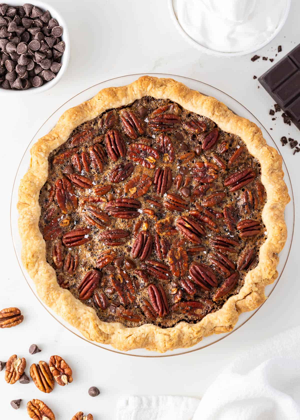 A luscious twist on a classic southern recipe, chocolate pecan pie is rich, velvety, and chocolatey loaded with pecans and topped with chocolate shavings and whipped cream. This is a decadent dessert you’ll want to show off! Chocolate pecan pie on a white counter.
