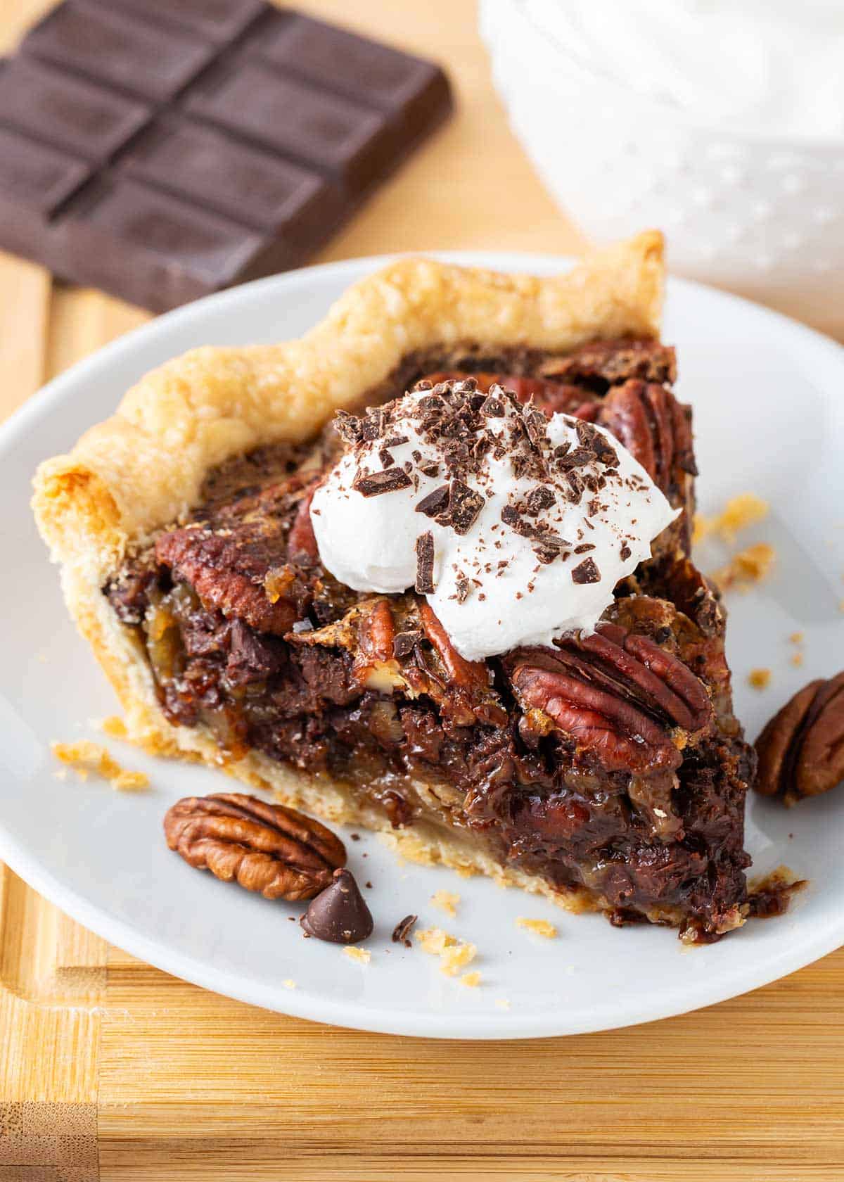 A luscious twist on a classic southern recipe, chocolate pecan pie is rich, velvety, and chocolatey loaded with pecans and topped with chocolate shavings and whipped cream. This is a decadent dessert you’ll want to show off! Slice of chocolate pecan pie on a white plate.