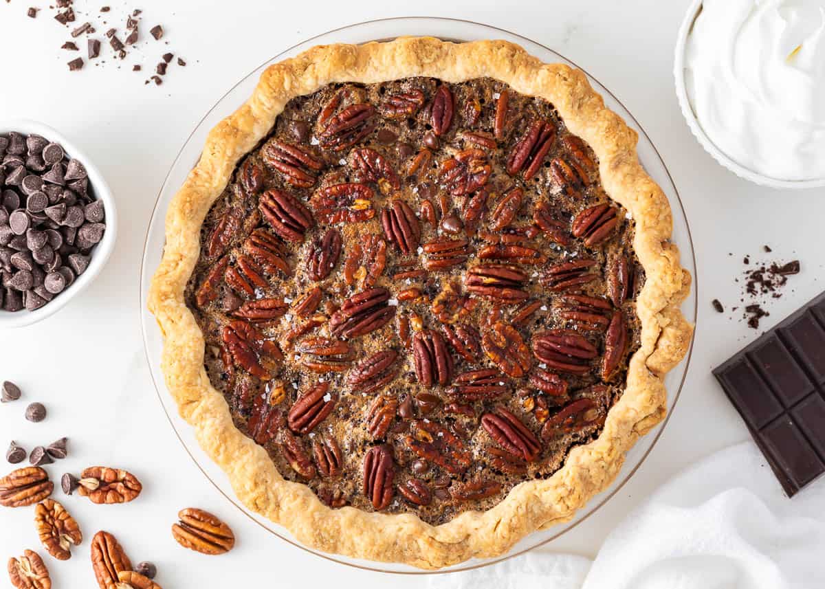 A luscious twist on a classic southern recipe, chocolate pecan pie is rich, velvety, and chocolatey loaded with pecans and topped with chocolate shavings and whipped cream. This is a decadent dessert you’ll want to show off! Chocolate pecan pie on a countertop.