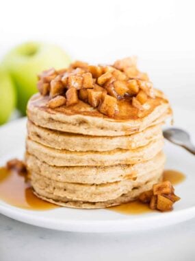 Tall stack of fluffy apple cinnamon pancakes topped with warm, diced cinnamon apples and drizzled with syrup, served on a white plate with a green apple in the background