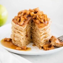 Plate of fluffy apple cinnamon pancakes with a wedge cut out, topped with warm cinnamon apples and syrup, with green apples in the background.