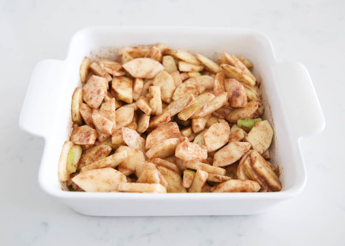 Sliced apples tossed with cinnamon and sugar, ready to be topped with crumble in a white baking dish.