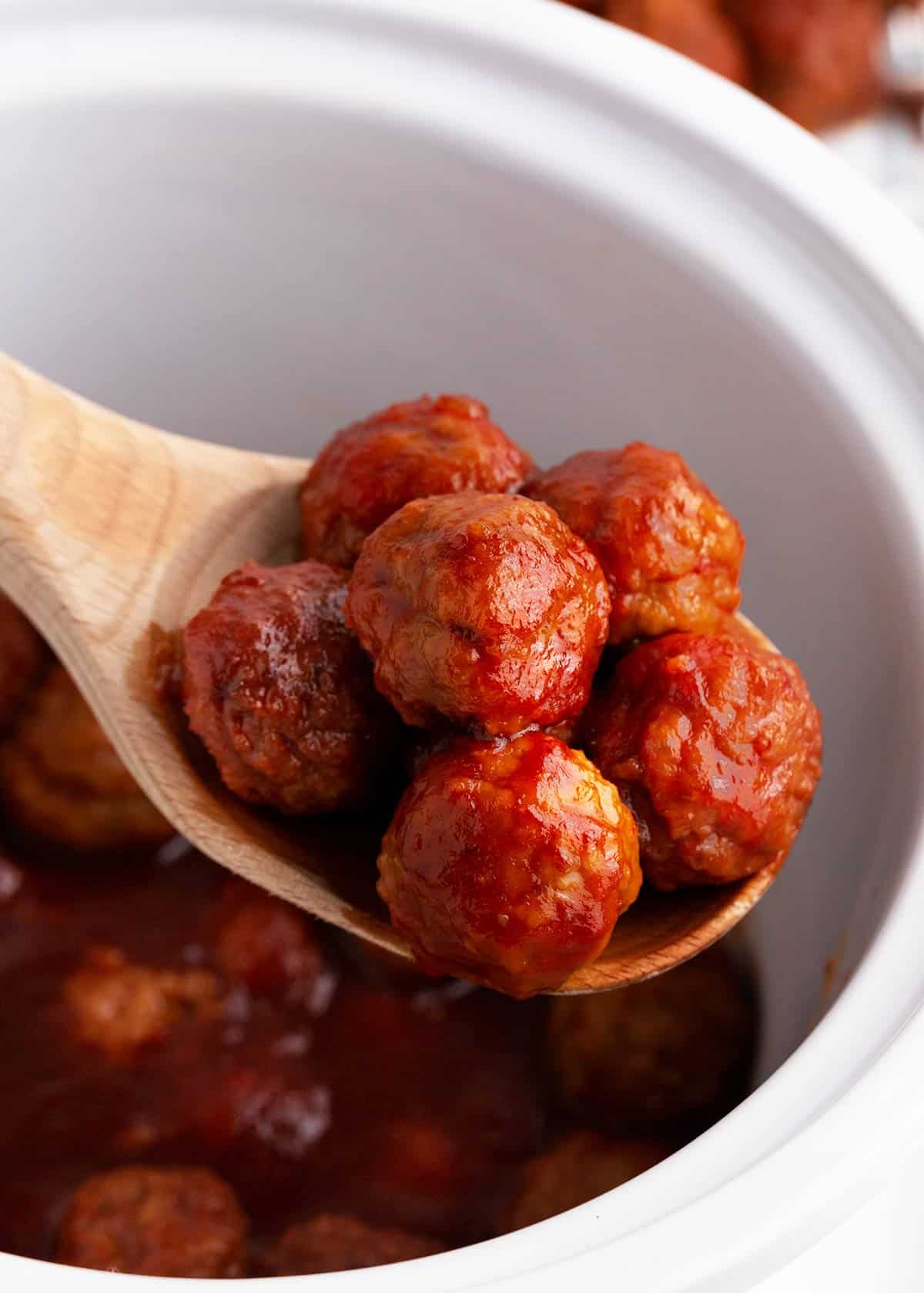 Meatballs being lifted out with a wooden spoon in a slow cooker. 