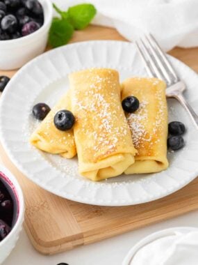 Cheese blintzes on a white plate dusted with powdered sugar and garnished with fresh blueberries.
