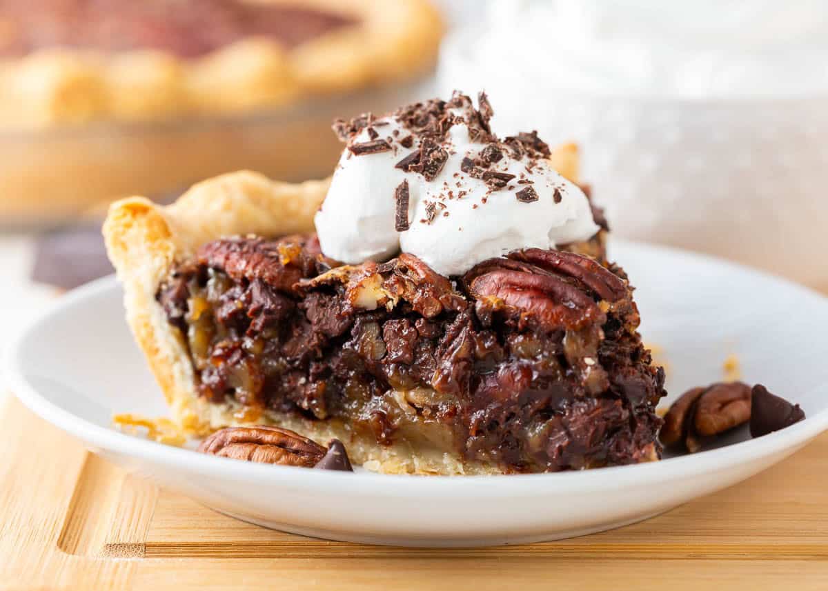 A luscious twist on a classic southern recipe, chocolate pecan pie is rich, velvety, and chocolatey loaded with pecans and topped with chocolate shavings and whipped cream. This is a decadent dessert you’ll want to show off! Slice of chocolate pecan pie on a white plate topped with whipped cream and chocolate shavings.