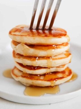 Tall stack of mini silver dollar pancakes on a white plate, drizzled with maple syrup and pierced with a fork.