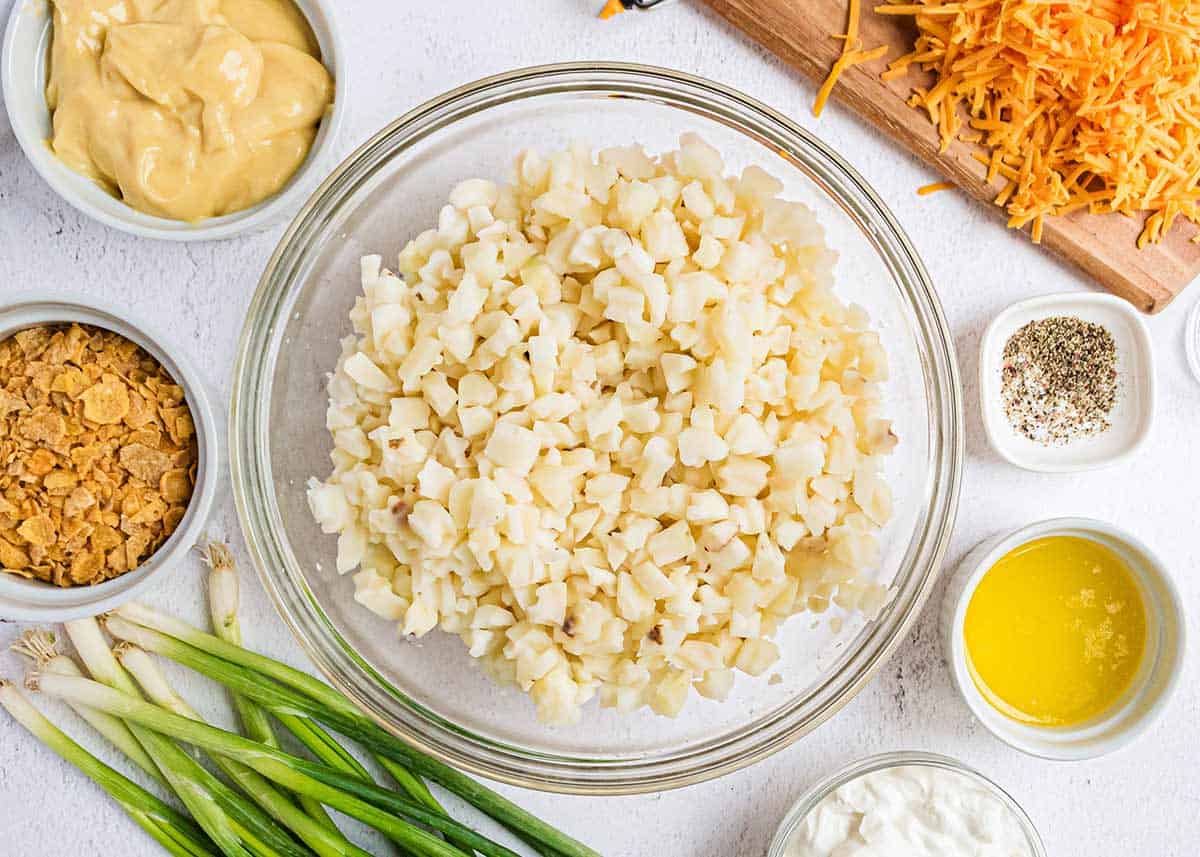 Diced potatoes in a bowl, with cornflakes, shredded cheddar cheese, cream of chicken soup, sour cream and butter on the counter for the casserole.