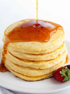 Fluffy homemade pancakes stacked on a plate with maple syrup pouring over the top.