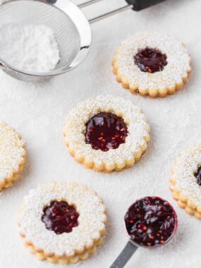 Linzer raspberry cookies on the counter with jam in a spoon and powdered sugar in a sifter.