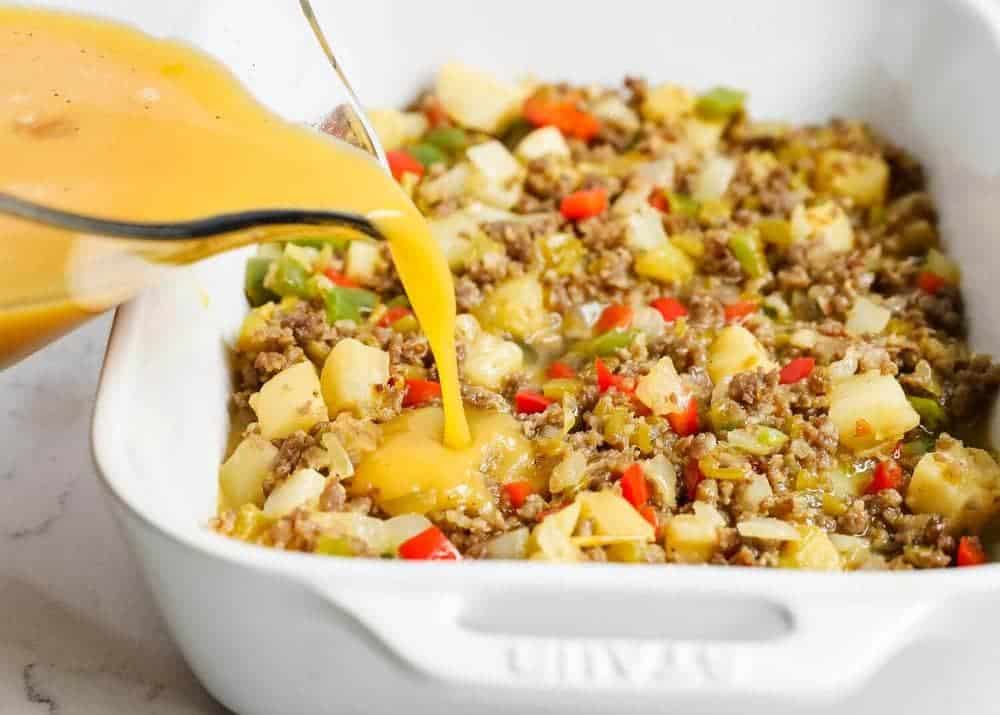 Whisked eggs being poured over sausage and potato mixture in a white baking dish.