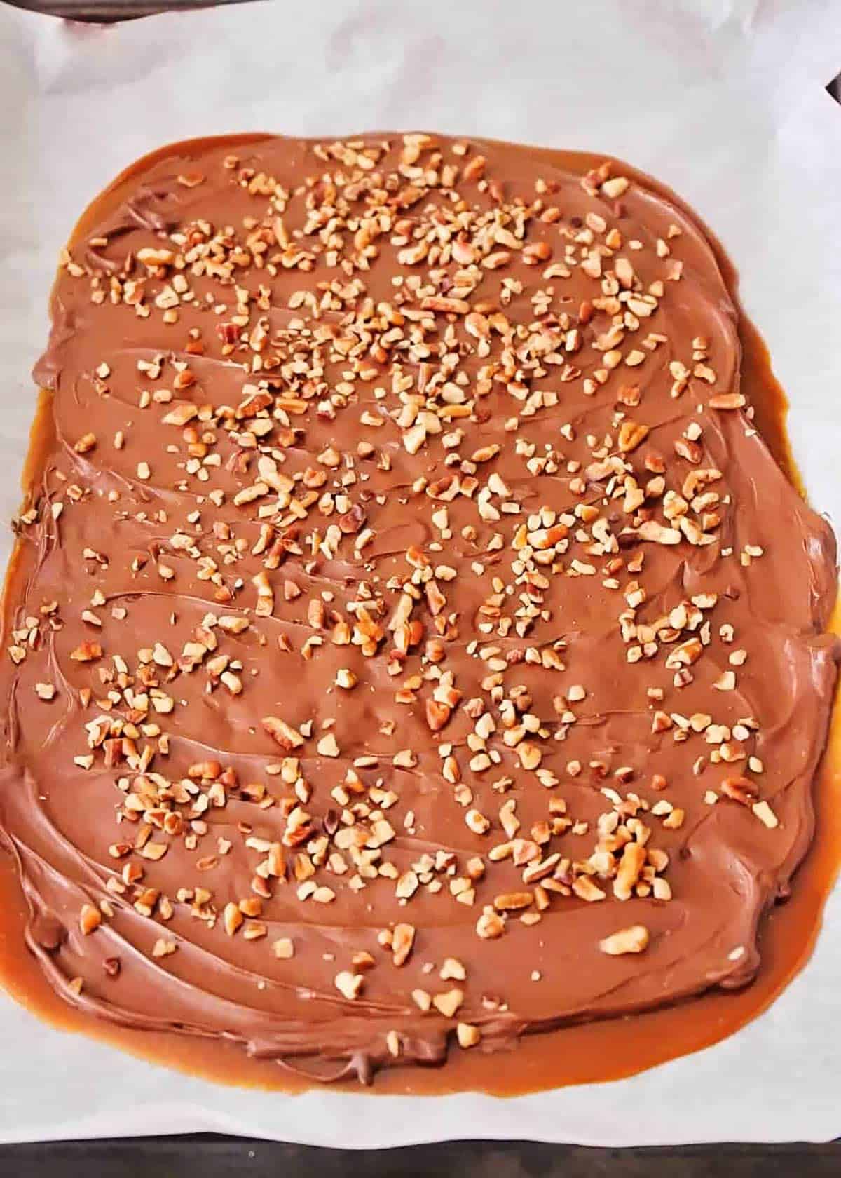 Finished toffee recipe spread out on parchment paper topped with chocolate and pecans.