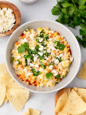 A bowl of elote dip with queso fresco cheese and cilantro surrounded by tortilla chips.