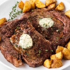 Oven baked steak with a pat of garlic butter on top with fresh herbs and roasted potatoes served on a plate.