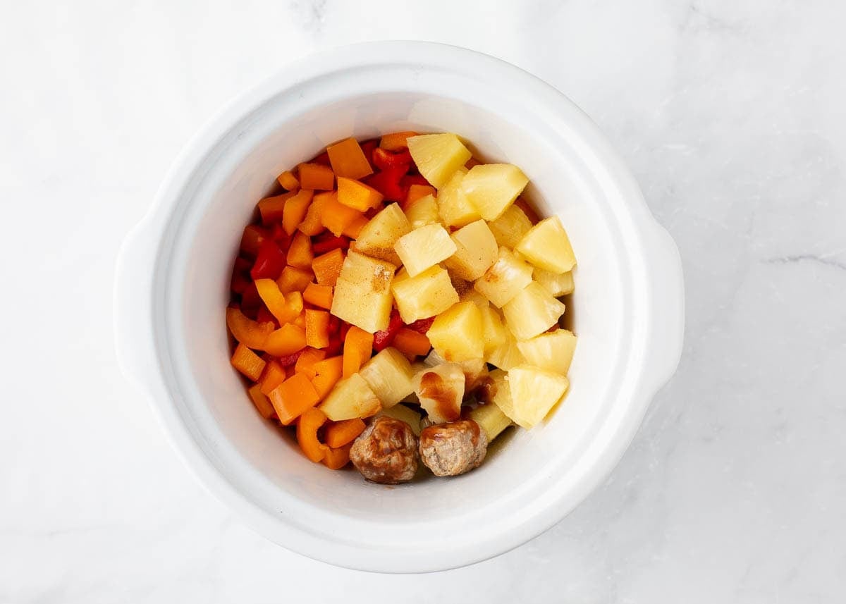 Frozen meatballs, pineapple chunks and diced bell peppers added to a white slow cooker before cooking