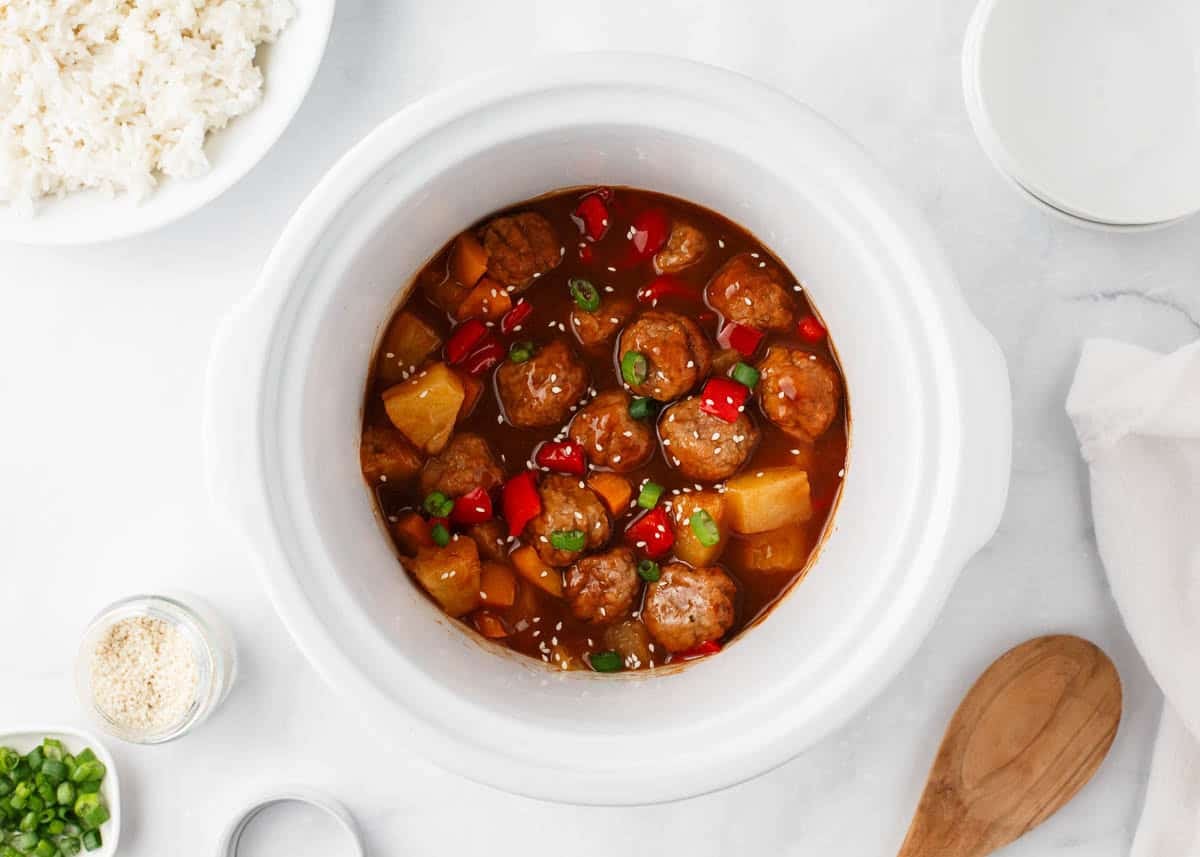 Hawaiian meatballs in a slow cooker with pineapple chunks, red bell pepper, green onions and sesame seeds.